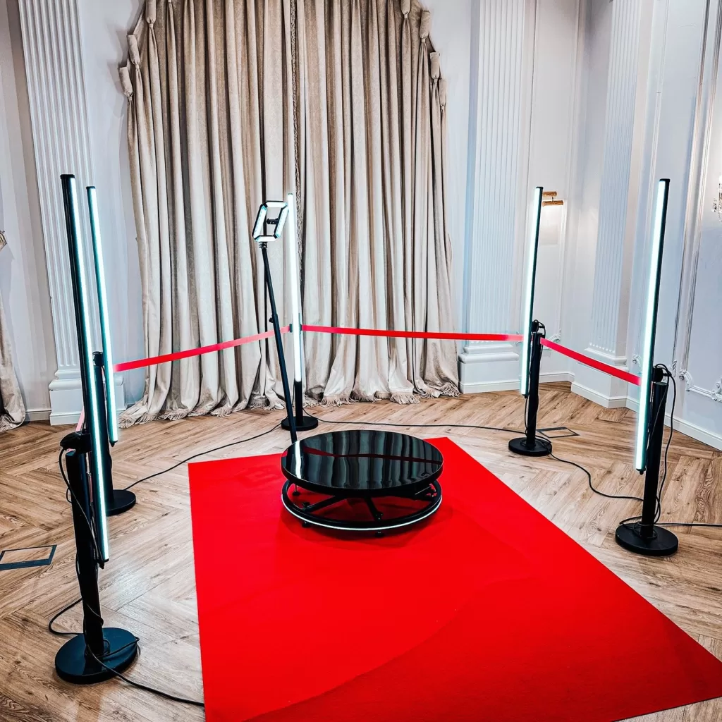 Photobooth 360 con base giratoria negra, alfombra roja y luces LED verticales, delimitado con cinta en un salón elegante.