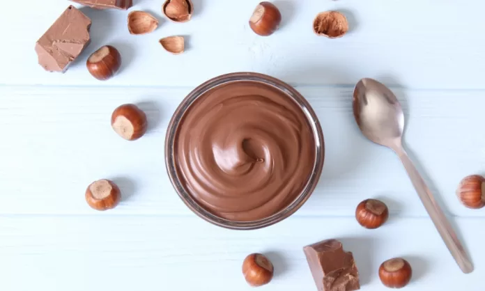 crema-de-avellanas3 Recipiente de vidrio con pasta cremosa de color chocolate rodeado de avellanas y trozos de tableta, sobre fondo claro