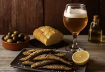 Boquerones: qué son, cómo se preparan y cómo servirlos en fiestas Boquerones fritos con limón servidos como tapa, acompañados de pan, aceitunas, aceite de oliva y cerveza sobre mesa rústica.