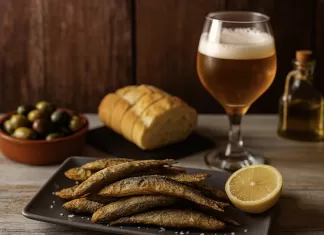 Boquerones: qué son, cómo se preparan y cómo servirlos en fiestas Boquerones fritos con limón servidos como tapa, acompañados de pan, aceitunas, aceite de oliva y cerveza sobre mesa rústica.