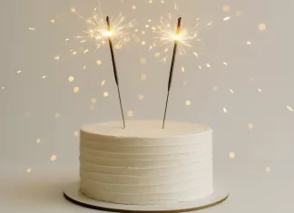 Velas de cumpleaños: tipos, tamaños y mejores opciones Pastel blanco minimalista con dos bengalas de cumpleaños encendidas y destellos dorados.