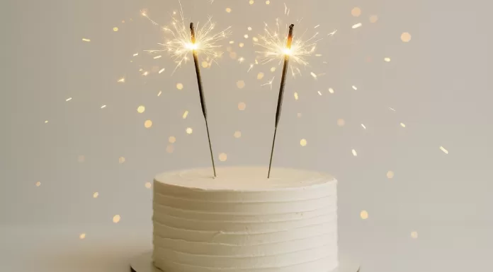 Velas de cumpleaños: tipos, tamaños y mejores opciones Pastel blanco minimalista con dos bengalas de cumpleaños encendidas y destellos dorados.