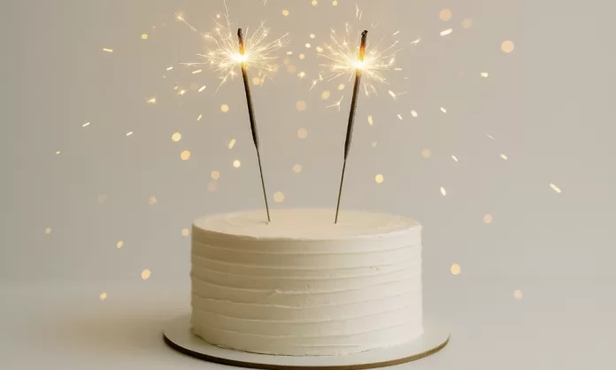 Pastel blanco minimalista con dos bengalas de cumpleaños encendidas y destellos dorados.